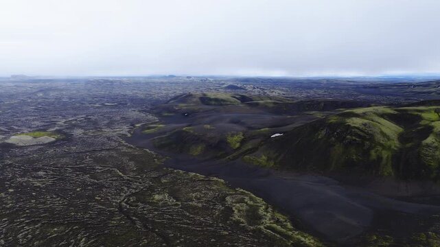 Aerial view captures the volcanic landscape of Laki in Iceland, featuring moss covered hills, black volcanic sand, and vast solidified lava fields beneath an overcast sky.