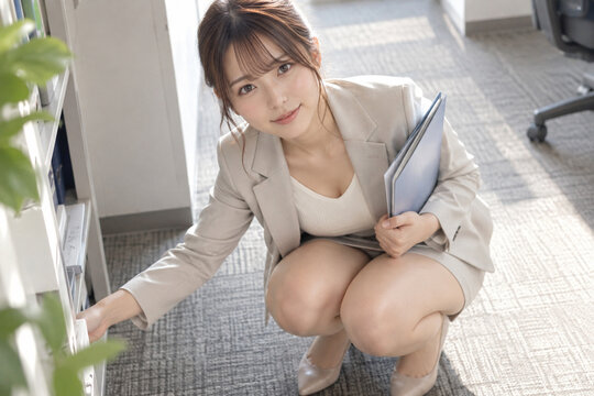 青いファイルを抱え白い棚に手を伸ばす淡色スーツ姿の女性
