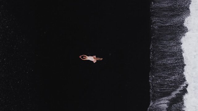 Aerial top down view of a beautiful woman in a dress lying on the dark volcanic black sand beach in Vik Iceland while ocean waves roll and foam onto the shore.