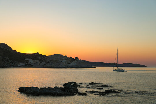 The silhouette of a sailboat during a beautiful sunset at Mandrakia on the island of Milos in Greece. The Greek sky is orange and red as the sun descends behind the rocky coastline. 
