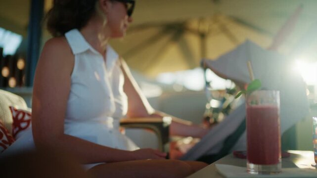 Mother in a white dress reaching for a drink on a sunlit lounge terrace beside a baby stroller, red cocktail in the foreground, French Riviera, France.