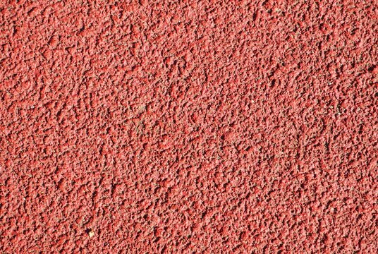 Close-up of a rough red granular surface with an irregular, densely packed texture and subtle variations in color and shadow. The detailed synthetic running track pattern fills the frame.