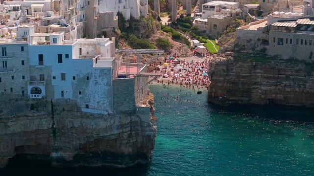 Polignano a Mare Puglia Italy aerial view of cliff houses, beach and Roman arch bridge. Close drone flight over historic town and Lama Monachile cove approaching ancient stone bridge 