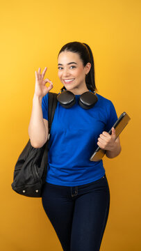 Happy latina student showing okay gesture while carrying a backpack and laptop