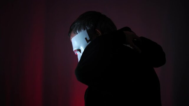 Mysterious person wearing a black hoodie and a mask for halloween or true crime against a dark red lit background