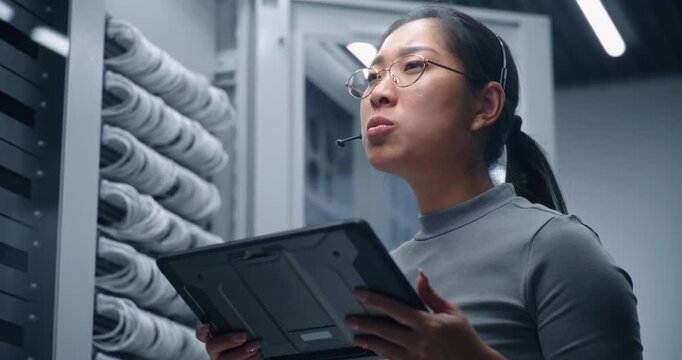 Female IT Specialist Reviews Connected Cables on Tablet While Speaking Through Headset. Technical Workflow Represents Network Inspection, Digital Reporting, Equipment Monitoring and Infrastructure