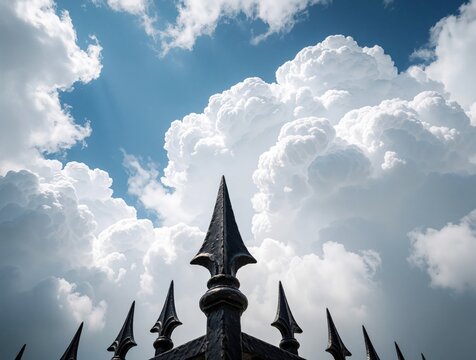 Close up of black iron fence with spear tips. Metal security gate stands against huge white cumulus clouds in blue sky. Sharp decorative spikes protect property boundary under bright sun. Vintage