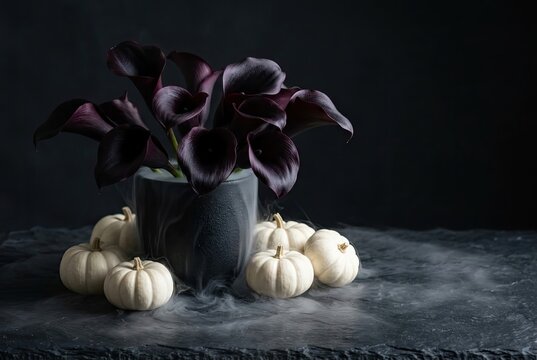 Calla lilies in a dark ceramic vase are surrounded by small white pumpkins sitting on a smoky slate surface.