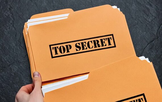 Top-down flat lay photo of a stack of orange manila file folders with white papers peeking out from the top edge, text "TOP SECRET" black rubber stamp mark on the front folder
