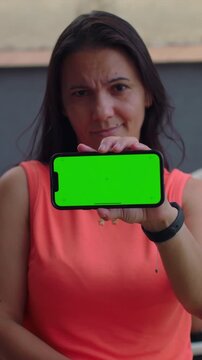 Woman holding smartphone green screen towards camera outdoors, neutral display mockup