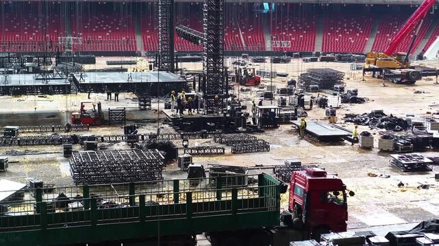 Cranes and construction crews assemble concert infrastructure inside a stadium lined with empty red seats.