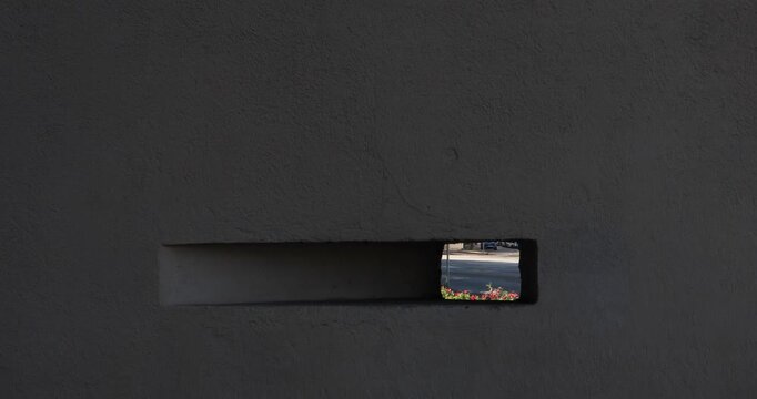 Dark concrete wall with narrow rectangular shaped opening revealing fragments of passing cars and street beyond, creating a minimalist study of concealment, perspective urban observation.