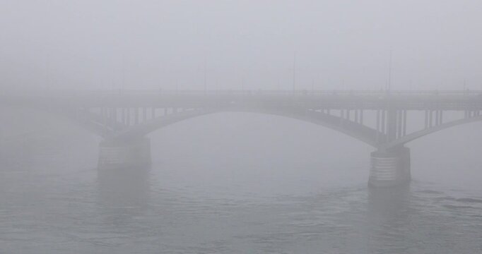 Basel&rsquo;s Wettsteinbr&uuml;cke emerges through dense fog above the Rhine, its steel arches and stone piers fading into a pale grey atmosphere with barely visible pedestrians and river ripples.