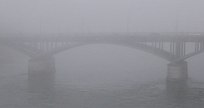 Basel&rsquo;s Wettsteinbr&uuml;cke emerges through dense fog above the Rhine, its steel arches and stone piers fading into a pale grey atmosphere with barely visible pedestrians and river ripples.