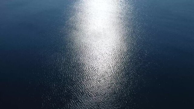 Aerial drone view of open sea with sunlight reflecting across the surface. Calm sea waves shimmering under direct sun rays during daylight.
