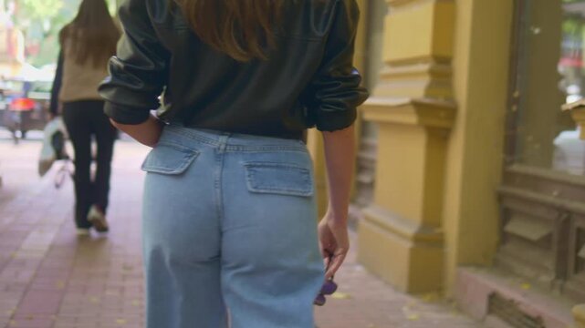 Woman walks away, jeans, leather jacket. Urban sidewalk, yellow building beside her. Her hand holds a small purse. Neutral expression, unaware of camera. Perfect for fashion, lifestyle, urban scenes
