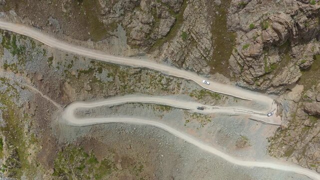 Top down drone view showing cars navigating sharp switchback turns on an unpaved mountain road cut into steep rocky terrain in Gilgit Baltistan Pakistan