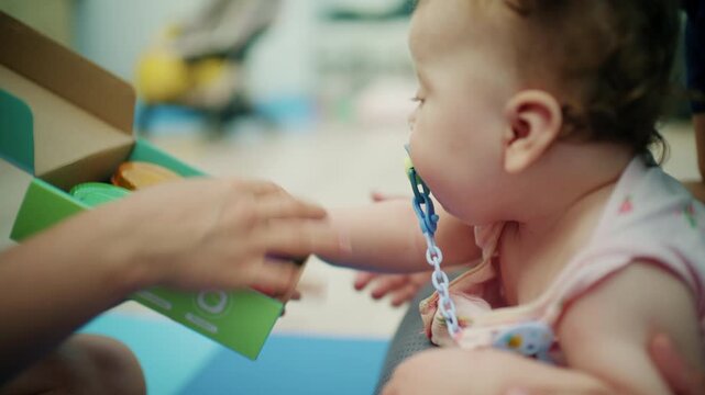 Curly-haired baby girl with a pacifier pressing colorful light-up buttons in a box held by a therapist during an early intervention session, French Riviera, France.