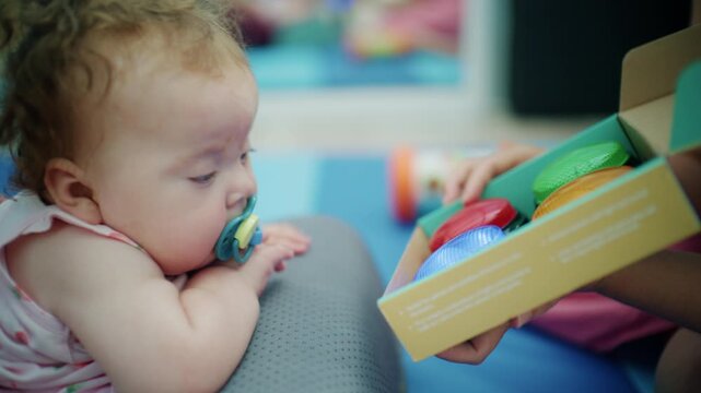 Baby girl in a pink romper leaning forward to touch red, green, blue and yellow buttons in a box while therapist hands guide her arm, French Riviera, France.