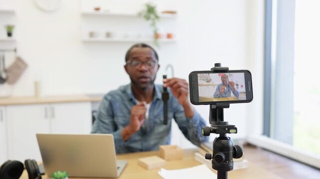 A bespectacled presenter gestures beside a laptop as a smartphone mounted on a tripod captures him in a bright, uncluttered kitchen