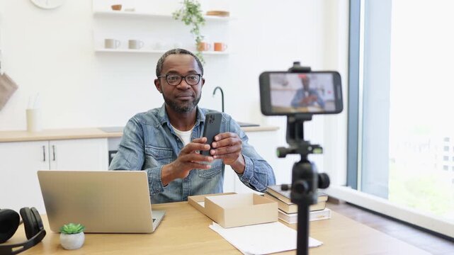 A creator demonstrates a smartphone at his kitchen table as a tripod-mounted camera captures him, with a laptop, open cardboard box, and papers nearby