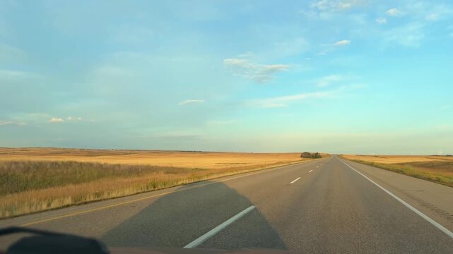 Driving down an empty straight highway through golden prairie fields in Saskatchewan under a blue sky, a road trip view across the open Canadian plains in summer.