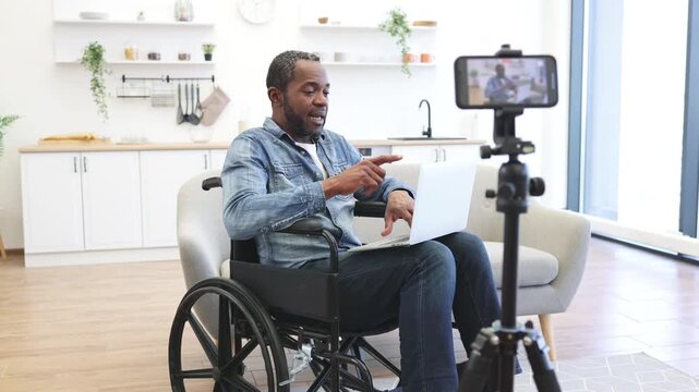 A seated creator gestures toward a camera while using a laptop in a bright home kitchen, with his wheelchair and recording setup clearly visible