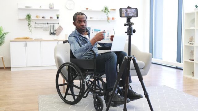 African American man in a wheelchair presents a printed chart beside his laptop, with a smartphone mounted on a tripod in a bright home interior