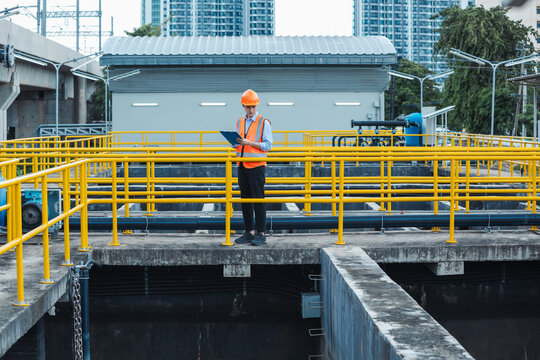 Young male engineer wearing vest and hard hat holding a clipboard inspecting wastewater in an industrial plant.