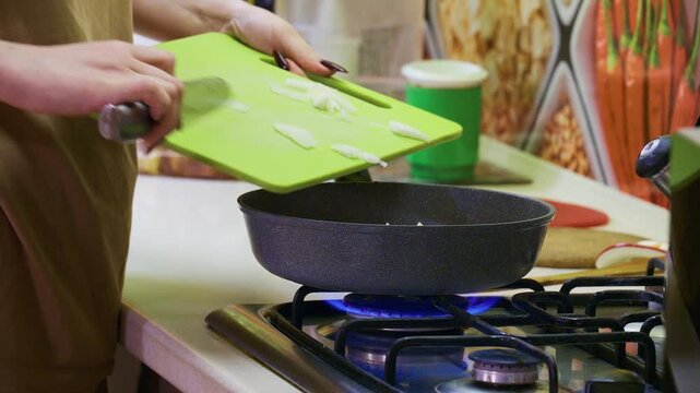Person slices onion on lime‑green cutting board; black frying pan sits on lit gas burner, green cup and kitchen backdrop visible.
