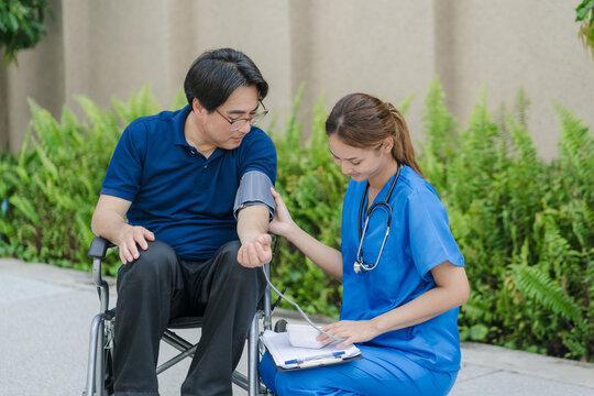 Asian female nurse measuring blood pressure of senior male patient in a wheelchair outdoor.