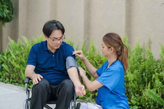 Asian female nurse measuring blood pressure of senior male patient in a wheelchair outdoor.