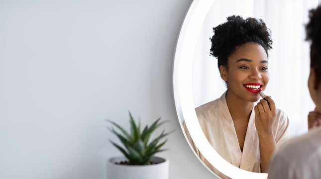 Young Black woman applying red lipstick looking at circular mirror with studio lighting