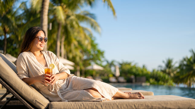 Young woman lounging on daybed at tropical pool resort holding cocktail