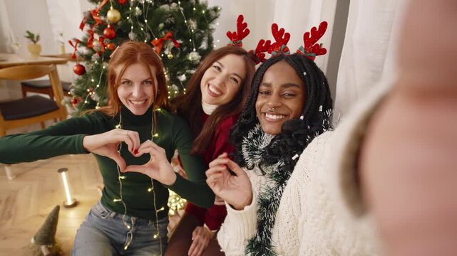 Three friends wearing reindeer antlers pose together make selfie beside a decorated Christmas tree indoors