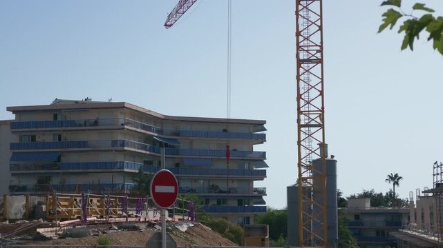 Construction site with a yellow tower crane, an apartment block with blue awnings, a no entry sign and a rising concrete building, French Riviera, France.