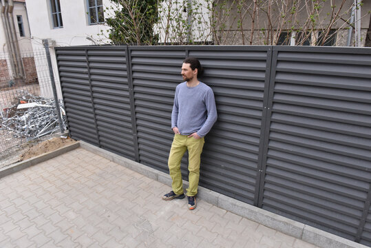 Man in grey sweater and yellow pants leaning against a dark louvered metal privacy fence