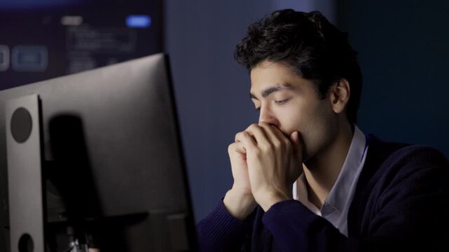 Computer Programmer considers difficult software problem beside multiple monitors, resting hands near face during concentrated technical work, reflecting challenging office computer work.