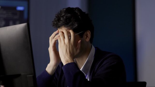 Software Developer covers face while seated before computer monitors, responding to challenging software issue within technology workplace, illustrating difficult computer-based activity.