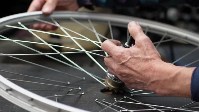Hands of a mechanic feeding spokes through the flange of a bicycle hub, wheel lying flat on a dark workbench. Lacing a wheel by hand, close up, shallow depth of field, real time 4K.