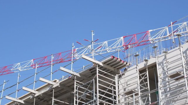 Red and white tower crane jib over the scaffolding and concrete walls of a new building under a clear blue sky, French Riviera, France.