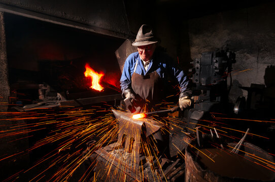 Senior blacksmith hitting glowing hot metal on anvil with shower of flying sparks in traditional workshop