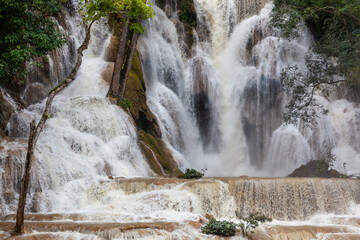 A wide waterfall cascades over rocky tiers into turquoise pools, framed by lush green trees in a dense forest at Kuang Si in Luang Prabang, Laos. © Eugene Ga