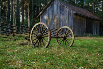 Antique wooden wagon wheels on an axle resting on green grass near a rustic fence and old wooden barn in the countryside © Dim