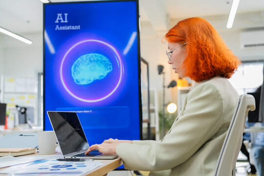 Redhead businesswoman in glasses typing on laptop and using AI assistant technology at office desk next to digital screen with artificial intelligence brain graphic