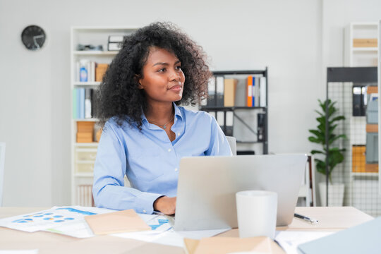 Thoughtful businesswoman with curly hair holding pen and looking at AI assistant screen while considering new technology as colleagues work in background office 