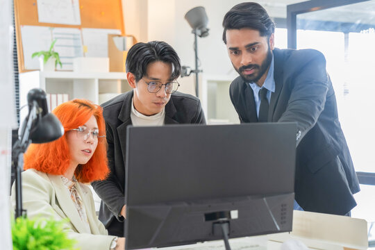 Bearded manager pointing at computer monitor while Asian and redhead colleagues focus on screen together, solving problem and reviewing project in modern office