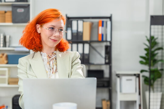 Smiling redhead businesswoman in glasses sitting at laptop and looking away with confident satisfied expression in bright modern office with shelves and plant 