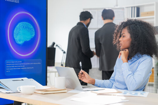 Thoughtful businesswoman with curly hair holding pen and looking at AI assistant screen while considering new technology as colleagues work in background office 