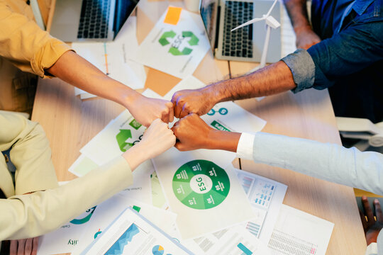 Top view of multiethnic team doing fist bump over ESG sustainability documents and wind turbine model, celebrating teamwork and green business success